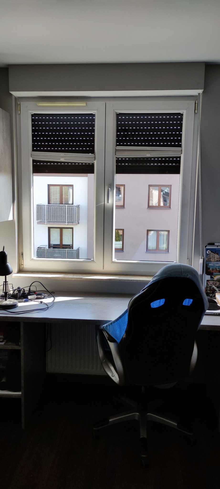 Pokój z biurkiem i krzesłem na tle okna z roletami, przez które widać budynek z balkonami. Na biurku lampka i akcesoria komputerowe.
