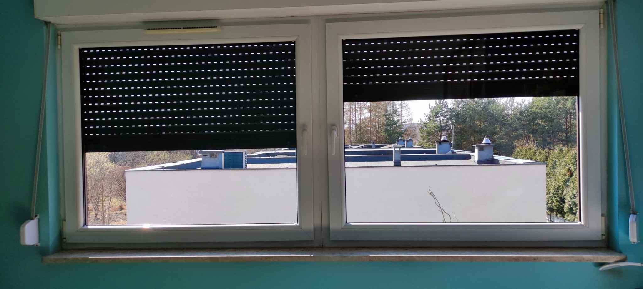 Białe okno PCV z częściowo zasłoniętymi roletami zewnętrznymi, przez które widać budynki i drzewa. Widok z wnętrza mieszkania na zewnątrz.