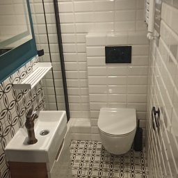 Paweł Sabiun - Wykończona łazienka z białą armaturą, mozaikową podłogą i białymi płytkami imitującymi cegłę. Umywalka z szafką z drewnopodobnego materiału. Kabina prysznicowa z czarną ramą.