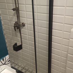 Paweł Sabiun - Nowoczesna kabina prysznicowa z czarnymi profilami i armaturą w kolorze starego złota, wykończona białą cegiełką i wzorzystą mozaiką podłogową.