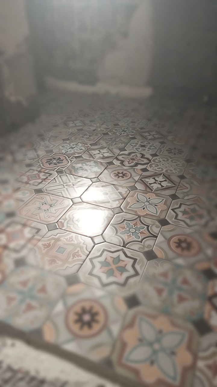 Ułożona podłoga z heksagonalnych płytek o wzorzystym, eklektycznym designie w odcieniach beżu, błękitu i szarości. W tle rozmyte, surowe ściany.