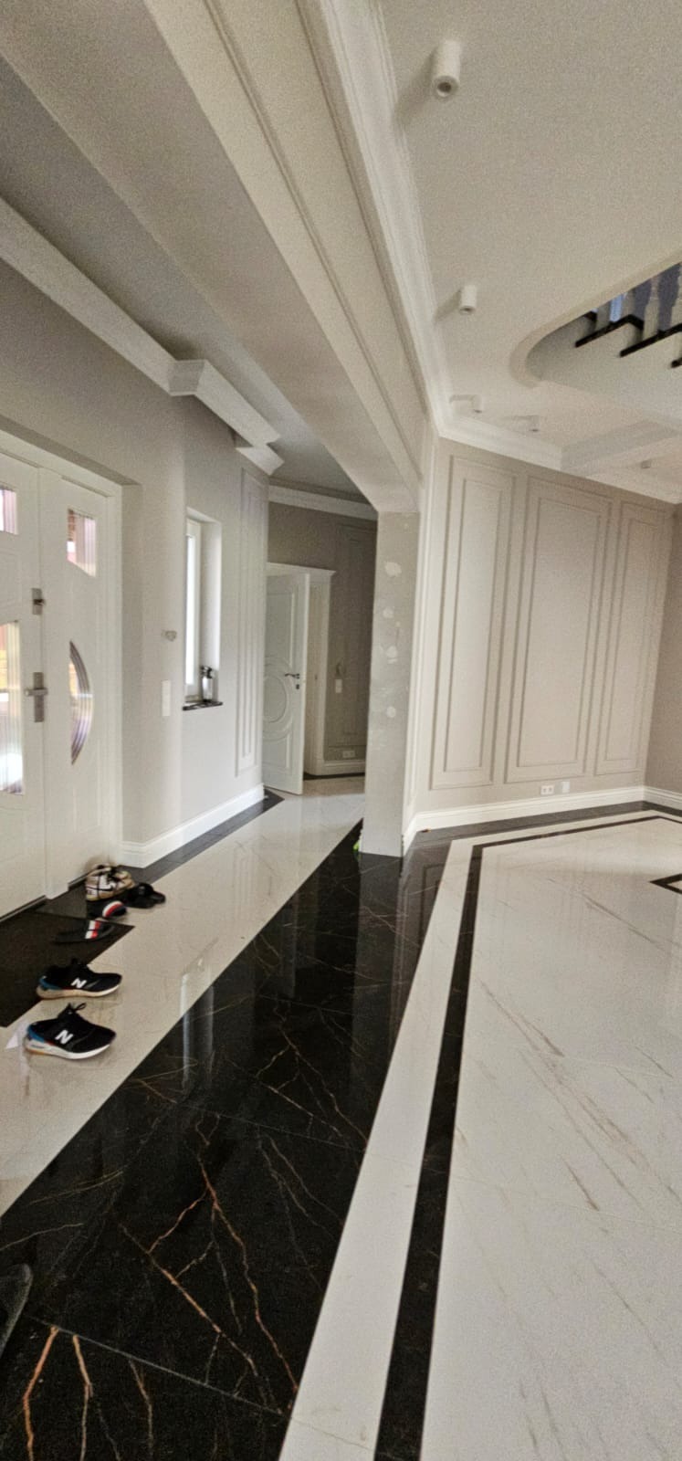 Elegancki hol z błyszczącą, czarno-białą marmurową podłogą w geometryczny wzór. Ściany w odcieniach szarości z dekoracyjnymi panelami i listwami.