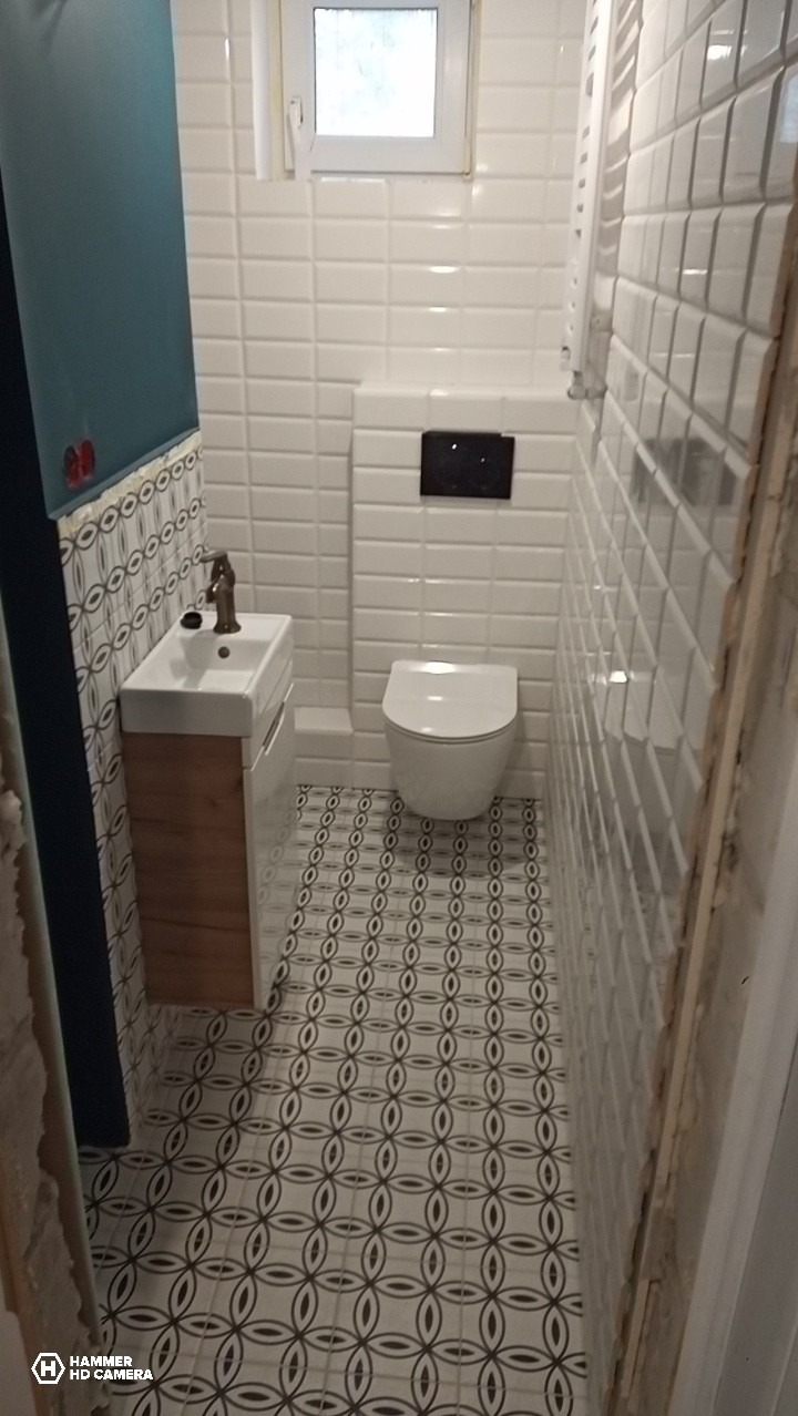 Wykończona łazienka z białą armaturą, mozaikową podłogą i ścianami z białych płytek. Umywalka z szafką w kolorze drewna. Wąskie pomieszczenie z oknem u góry.