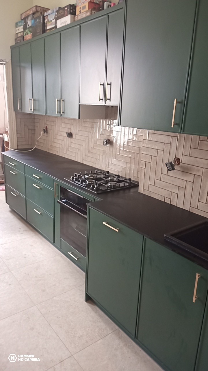 Nowoczesna kuchnia w odcieniach zieleni z czarnym blatem i płytkami ułożonymi w jodełkę. Nad szafkami widoczne pudełka gier planszowych.