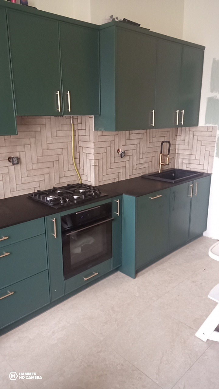 Zrealizowana kuchnia w odcieniach zieleni z płytkami ułożonymi w jodełkę na ścianie. Nowoczesne wykończenie z czarnym blatem i złotymi akcentami.