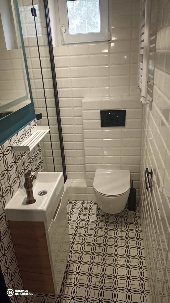 Wykończona łazienka z białą armaturą, mozaikową podłogą i białymi płytkami imitującymi cegłę. Umywalka z szafką z drewnopodobnego materiału. Kabina prysznicowa z czarną ramą.