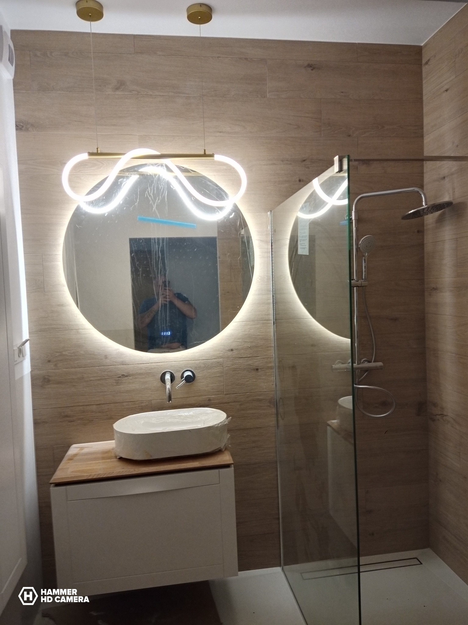 Nowoczesna łazienka z kabiną prysznicową, okrągłym lustrem z podświetleniem LED i umywalką na szafce. Płytki imitujące drewno na ścianach. Widoczne odbicie osoby w lustrze.