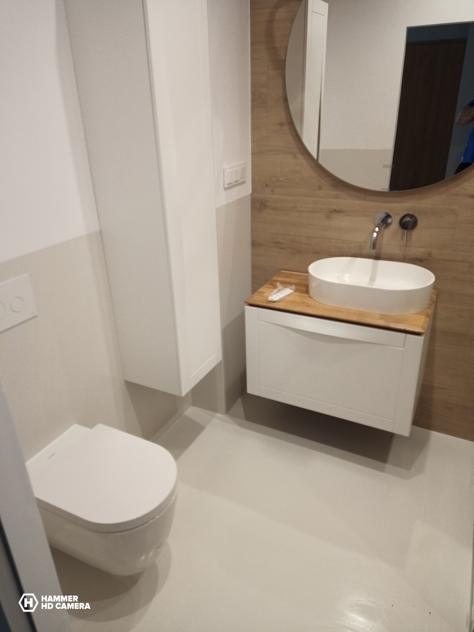 Nowoczesna łazienka po remoncie: biała toaleta, szafka z umywalką na drewnianym blacie i okrągłe lustro na ścianie wyłożonej płytkami imitującymi drewno.