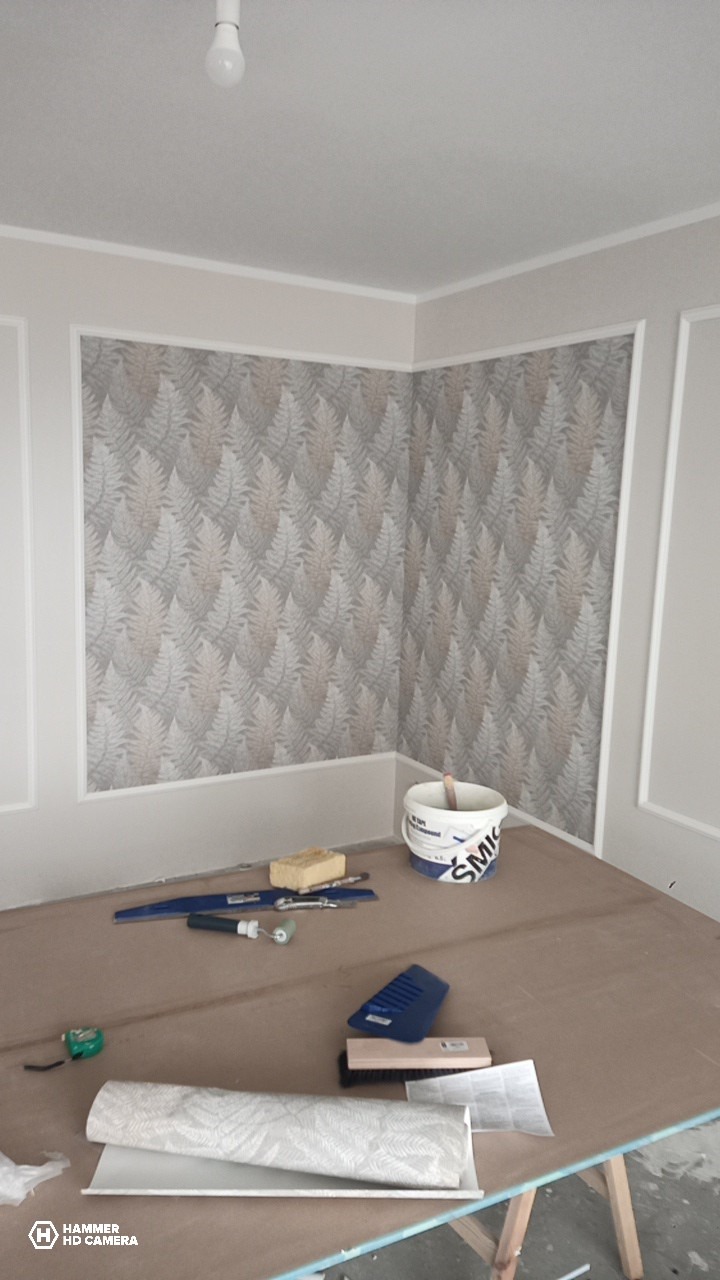 Tapetowanie ścian w pokoju z dekoracyjną sztukaterią. Widoczne narzędzia, wiadro z klejem i rolka tapety w roślinny wzór. Jasne wnętrze w trakcie prac wykończeniowych.