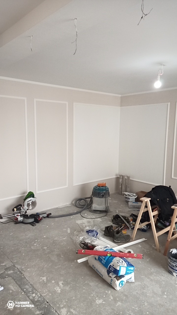 Pokój w trakcie wykańczania: listwy dekoracyjne na ścianach, narzędzia i materiały budowlane na podłodze. Widoczne przewody wystające z sufitu.