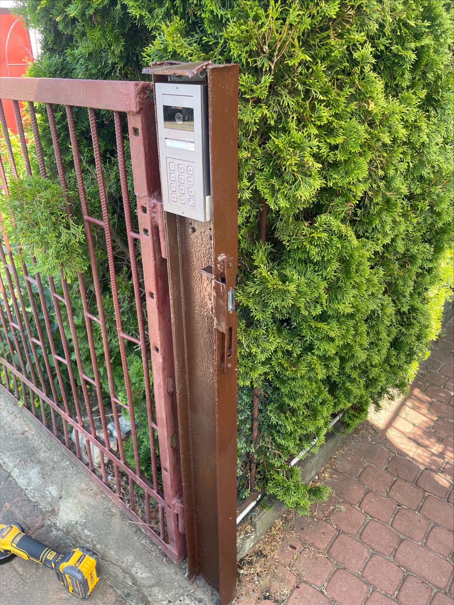 Brązowa furtka z domofonem przy zielonym żywopłocie, widoczna z bliska. Na dole leży szlifierka kątowa. Ujęcie z poziomu gruntu, w tle kostka brukowa.