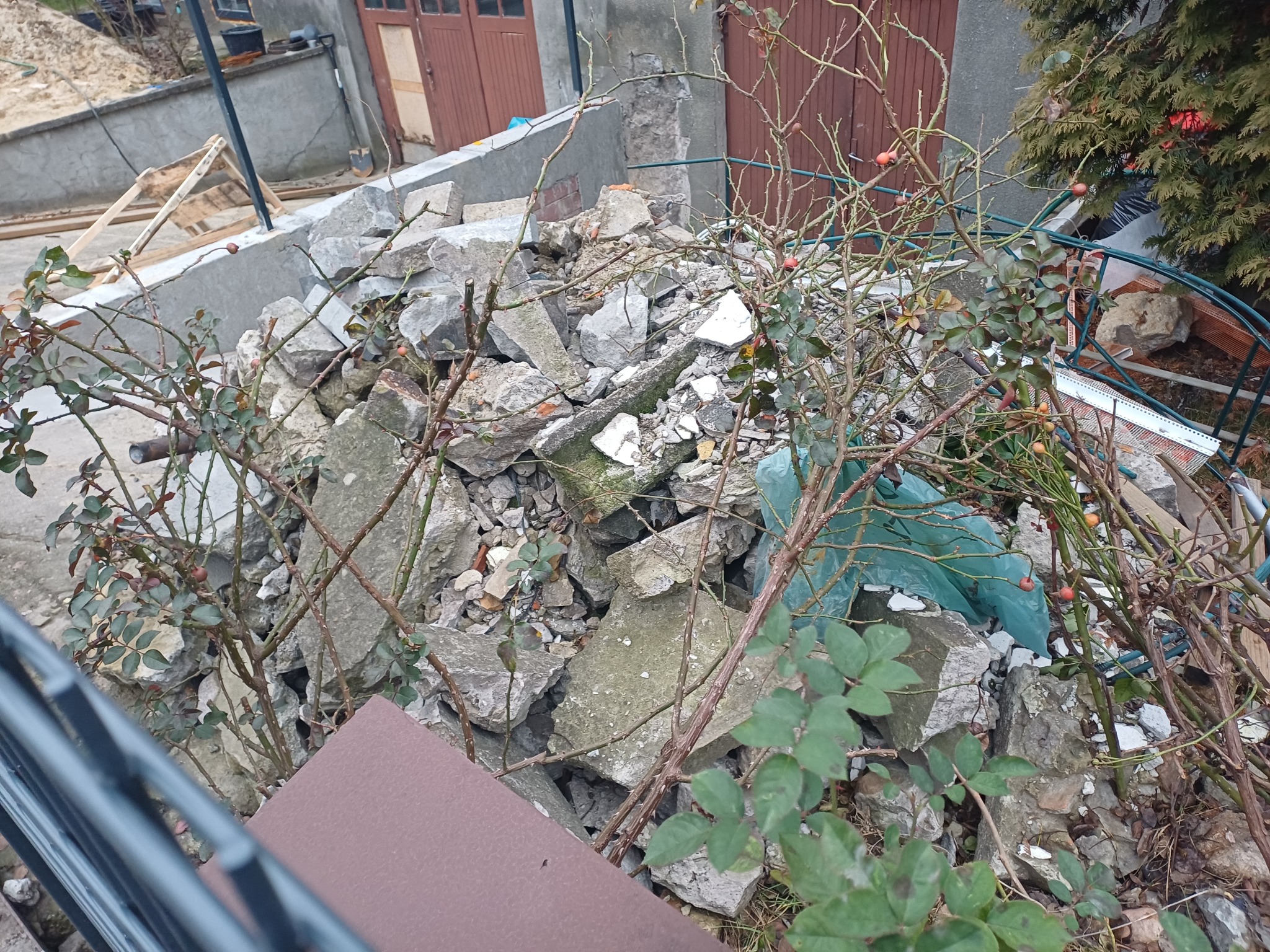 Sterta gruzu i odpadów budowlanych po remoncie, częściowo zasłonięta przez gałęzie krzewów. W tle widoczne drzwi garażowe i fragment ściany budynku.