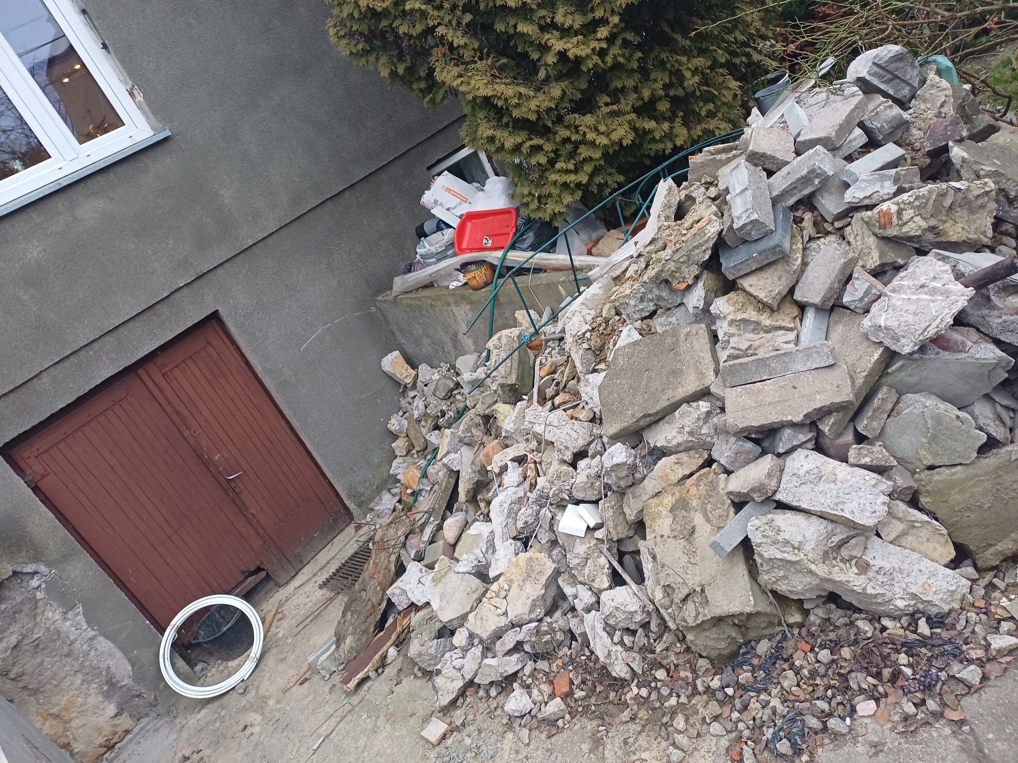 Sterta gruzu i odpadów budowlanych przy ścianie budynku z brązowymi drzwiami. Widoczne elementy wyposażenia wnętrz i zielone krzewy w tle.