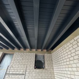 Usługi Ogólnobudowlane Sebastian Chyrzyński - Pomieszczenie w trakcie prac wykończeniowych: surowa cegła, okna zabezpieczone folią, pomalowany na ciemno drewniany strop. Widoczna drabina.