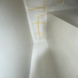 Usługi Ogólnobudowlane Sebastian Chyrzyński - Przygotowanie rogu ściany i sufitu do malowania: zabezpieczenie folią i taśmą malarską. Widok z dołu, jasne wnętrze, ostre światło.