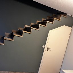 Usługi Ogólnobudowlane Sebastian Chyrzyński - Szare ściany z drewnianymi schodami i białymi drzwiami. Na pierwszym stopniu butelka. Minimalistyczny design wnętrza z ciemną górną częścią ściany.
