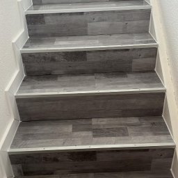 Usługi Ogólnobudowlane Sebastian Chyrzyński - Schody wykończone panelami w odcieniach szarości, z białymi listwami przypodłogowymi i metalowymi profilami na krawędziach stopni. Widok z dołu, oświetlenie naturalne.