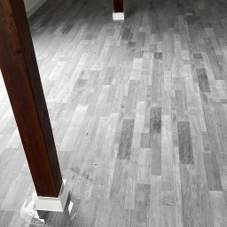 Usługi Ogólnobudowlane Sebastian Chyrzyński - Szare panele podłogowe ułożone w jodełkę francuską, z widocznymi drewnianymi słupami konstrukcyjnymi. Minimalistyczne wnętrze poddasza z białymi ścianami.