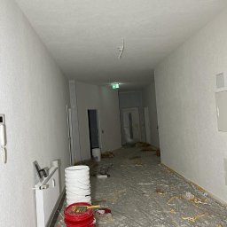 Usługi Ogólnobudowlane Sebastian Chyrzyński - Biały korytarz po świeżym malowaniu, widoczne wiadra z farbą, folia ochronna na podłodze i narzędzia malarskie. Ściany gładkie, jednolite, z widocznym grzejnikiem.