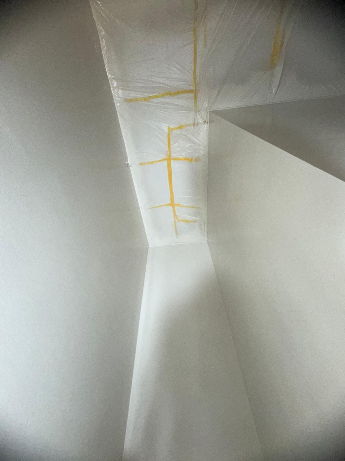 Przygotowanie rogu ściany i sufitu do malowania: zabezpieczenie folią i taśmą malarską. Widok z dołu, jasne wnętrze, ostre światło.