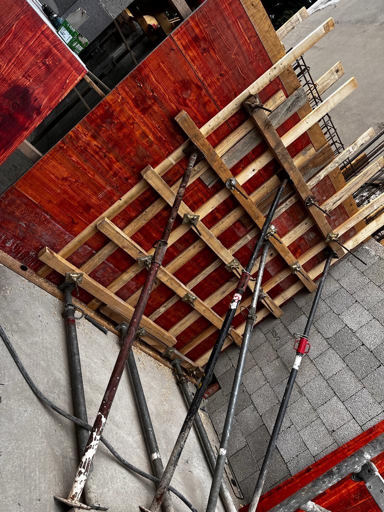 Szczegółowe ujęcie szalunku stropowego z czerwonymi deskami i stalowymi podporami, widoczne zbrojenie, na tle szarej powierzchni.