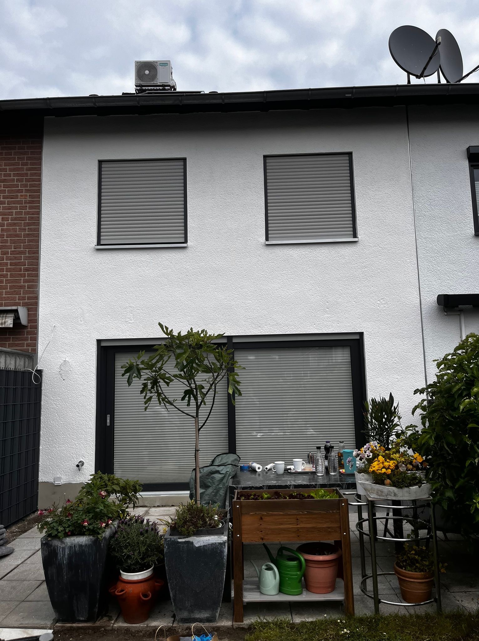 Biała elewacja domu z oknami z żaluzjami, dekorowana roślinami w donicach i stolikiem z akcesoriami ogrodowymi. Widoczny fragment ceglanej ściany i klimatyzator na dachu.