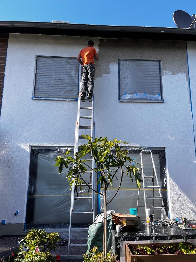 Pracownik na drabinie maluje elewację domu. Okna zabezpieczone folią. Widoczne narzędzia i roślina w donicy. Częściowo pomalowana ściana w jasnym odcieniu.