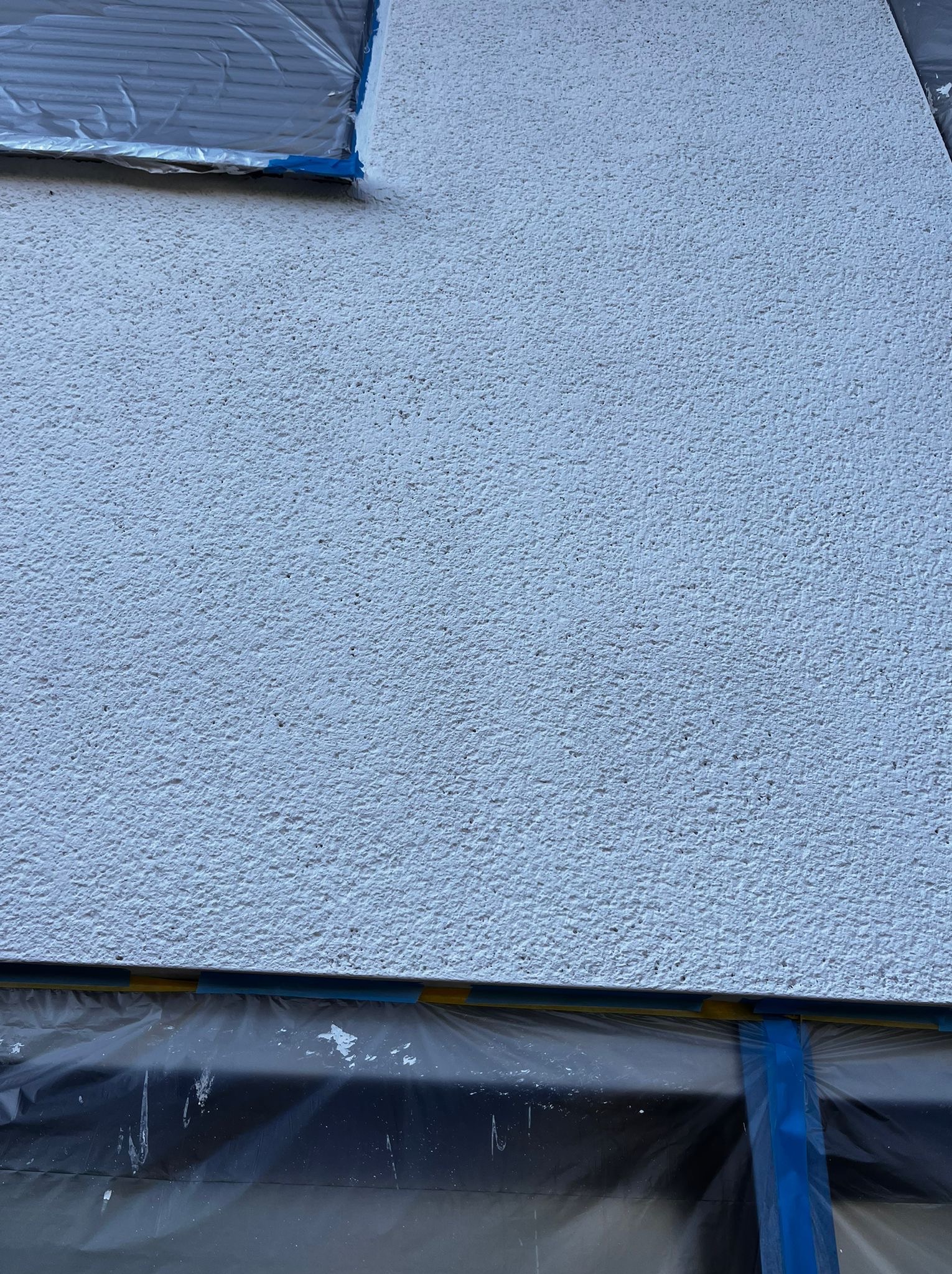 Biała, chropowata elewacja po nałożeniu tynku baranka, zabezpieczona folią malarską przy oknie i cokole. Widoczne detale faktury i staranne zabezpieczenie.