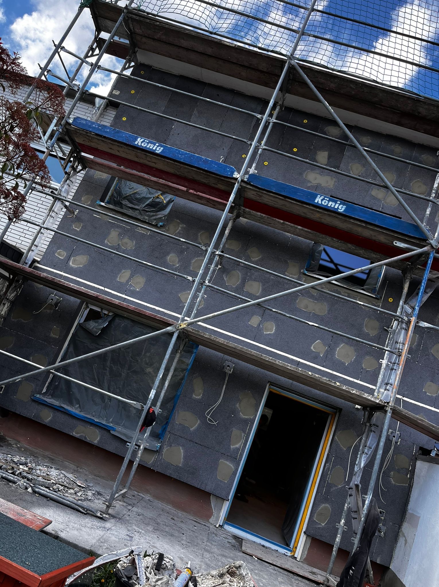 Ocieplanie elewacji budynku z użyciem rusztowania. Widoczne płyty izolacyjne z wypełnionymi otworami. Okna zabezpieczone folią. Ujęcie z dołu, pochylone.