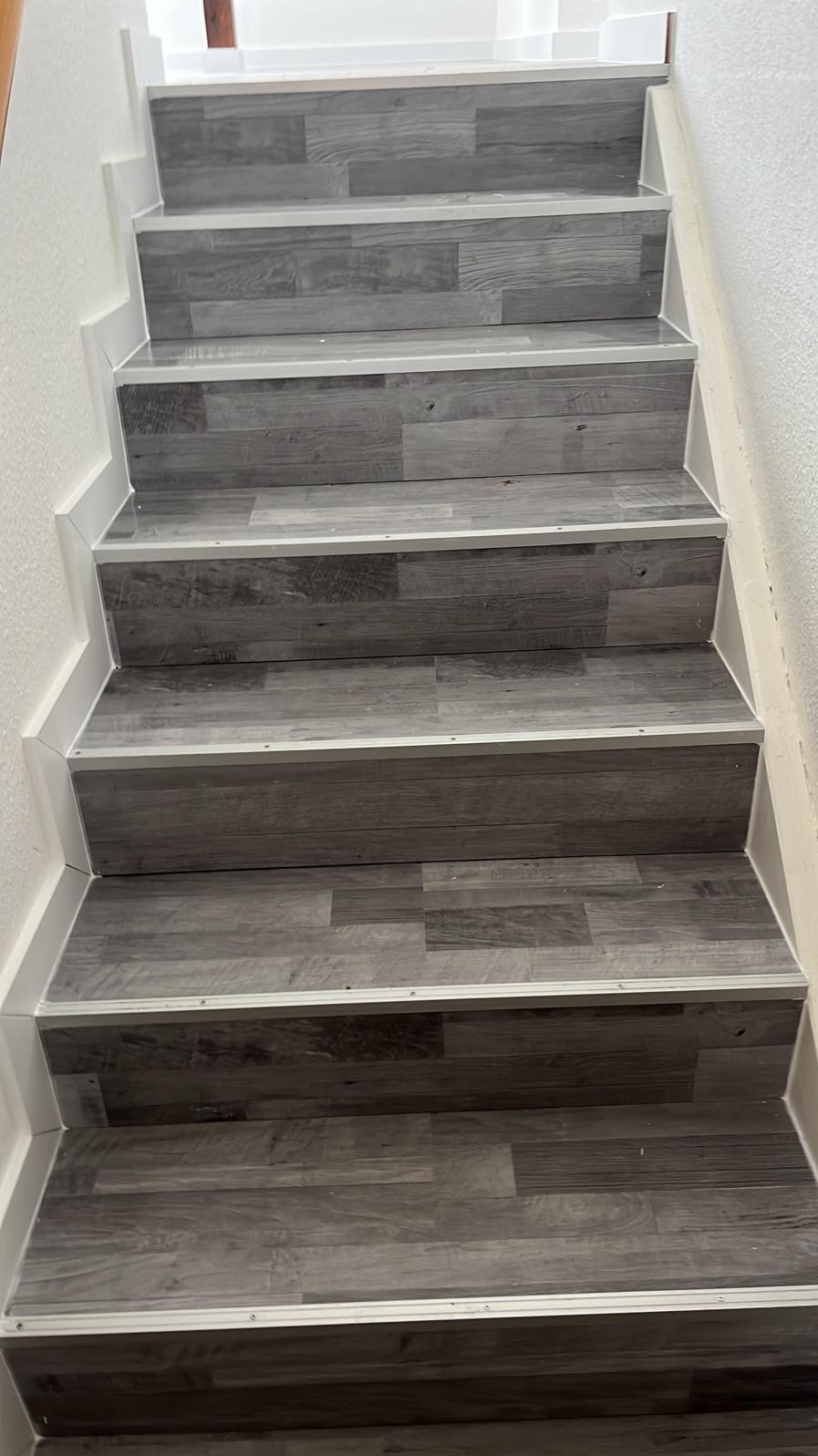 Schody wykończone panelami w odcieniach szarości, z białymi listwami przypodłogowymi i metalowymi profilami na krawędziach stopni. Widok z dołu, oświetlenie naturalne.