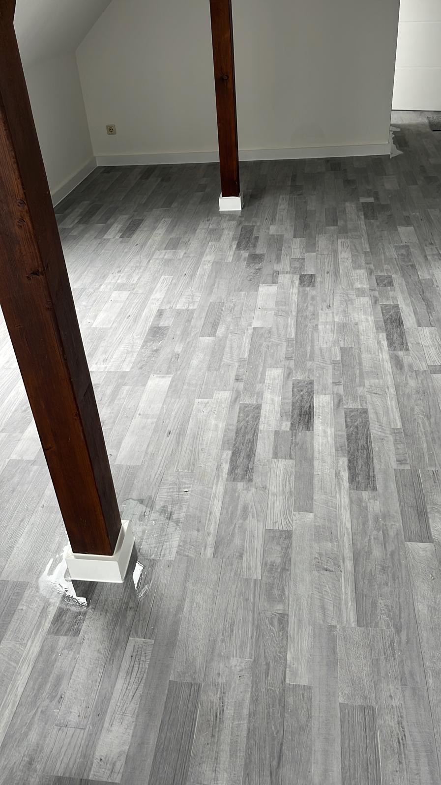 Szare panele podłogowe ułożone w jodełkę francuską, z widocznymi drewnianymi słupami konstrukcyjnymi. Minimalistyczne wnętrze poddasza z białymi ścianami.