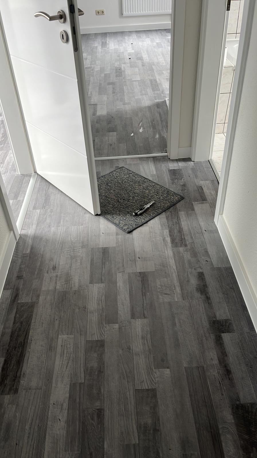 Ułożone szare panele podłogowe imitujące drewno, widok z perspektywy wejścia do pomieszczenia, z widoczną wycieraczką i nożykiem do paneli.