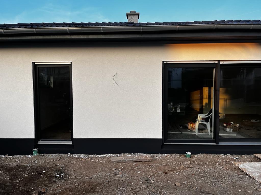 Biała elewacja domu z czarnymi ramami okiennymi i podmurówką. Widoczne ślady po instalacjach na ścianie. Teren wokół domu częściowo przygotowany.