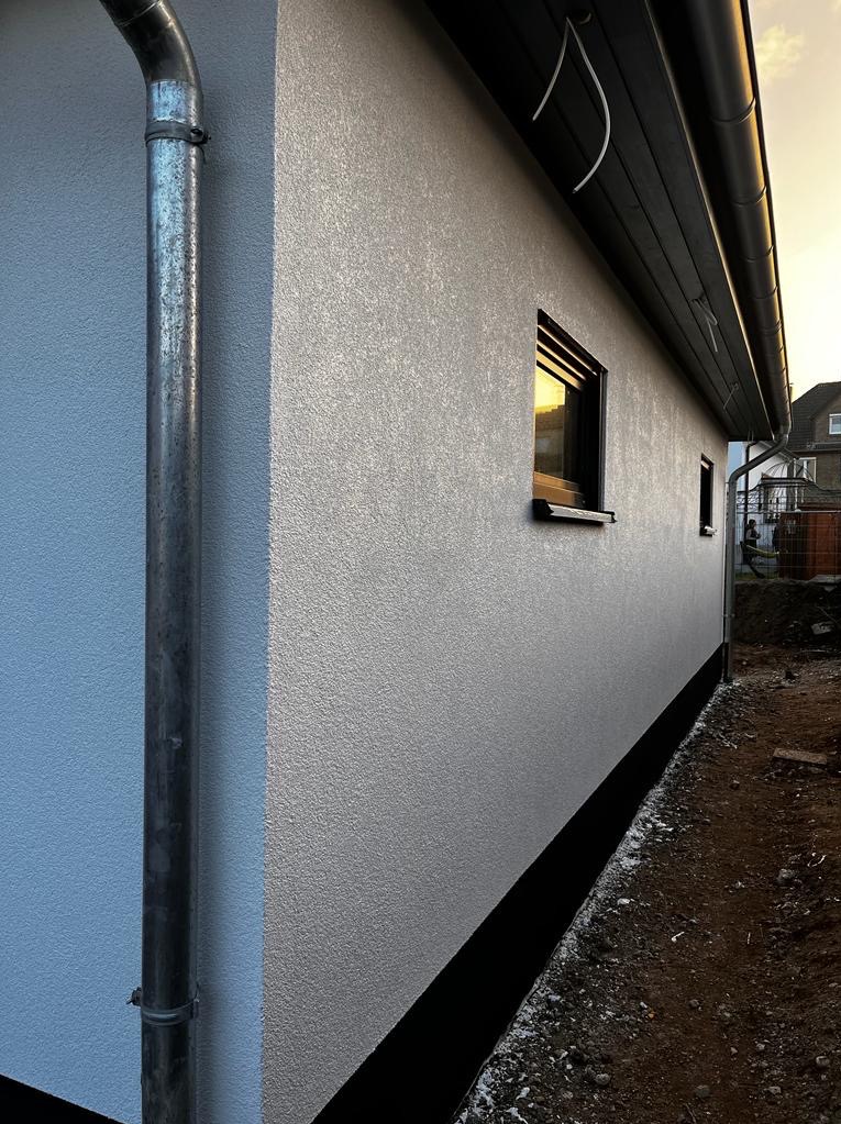 Ściana budynku po nałożeniu strukturalnego tynku w odcieniach szarości, z widoczną rynną i oknem, w którym odbija się zachodzące słońce. Ujęcie z boku budynku.
