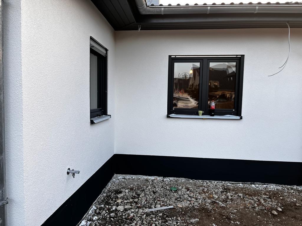 Biała elewacja budynku z dwoma czarnymi oknami i czarnym pasem u dołu. Widoczne kamienie na ziemi i rynna. Na parapecie stoi napój i kubek.