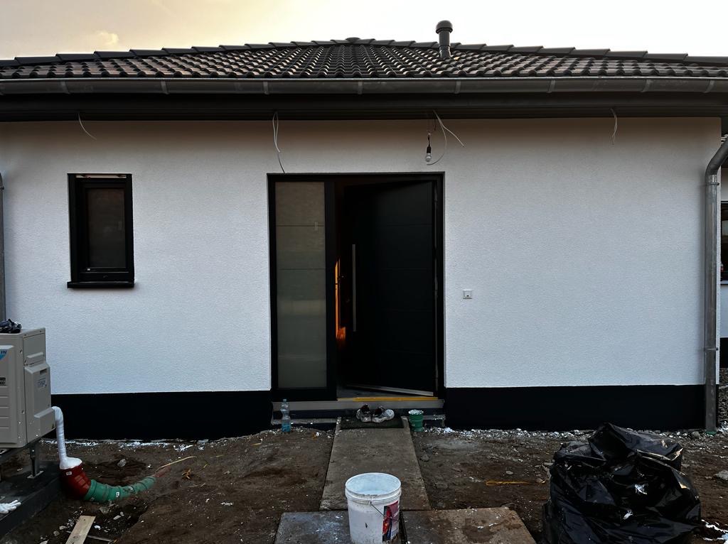 Biała elewacja domu jednorodzinnego z czarnymi detalami: okno, drzwi wejściowe i cokół. Widoczne ślady prac wykończeniowych wokół budynku. Otwarta nowoczesna stolarka drzwiowa.