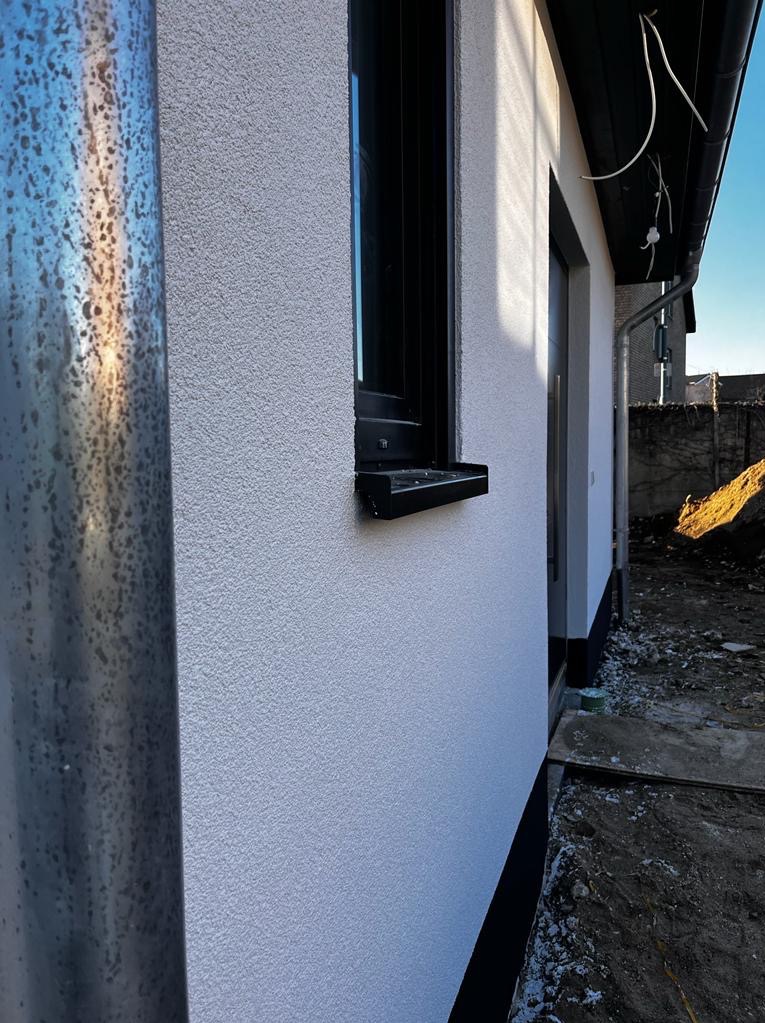 Biała elewacja domu z czarnymi detalami: okno z parapetem i fragment rynny. Widoczna faktura tynku i fragment otoczenia budynku w słoneczny dzień.