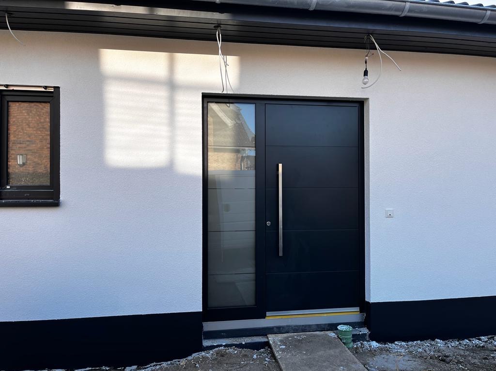 Nowoczesne, czarne drzwi wejściowe z długim, srebrnym uchwytem, wkomponowane w białą elewację budynku z czarnym cokołem i małym oknem po lewej stronie.