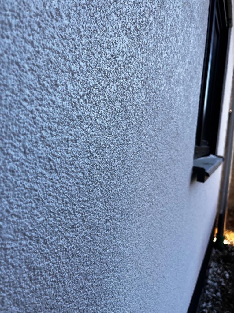 Szara, ziarnista faktura elewacji budynku z widocznym fragmentem okna. Delikatne światło podkreśla strukturę tynku.