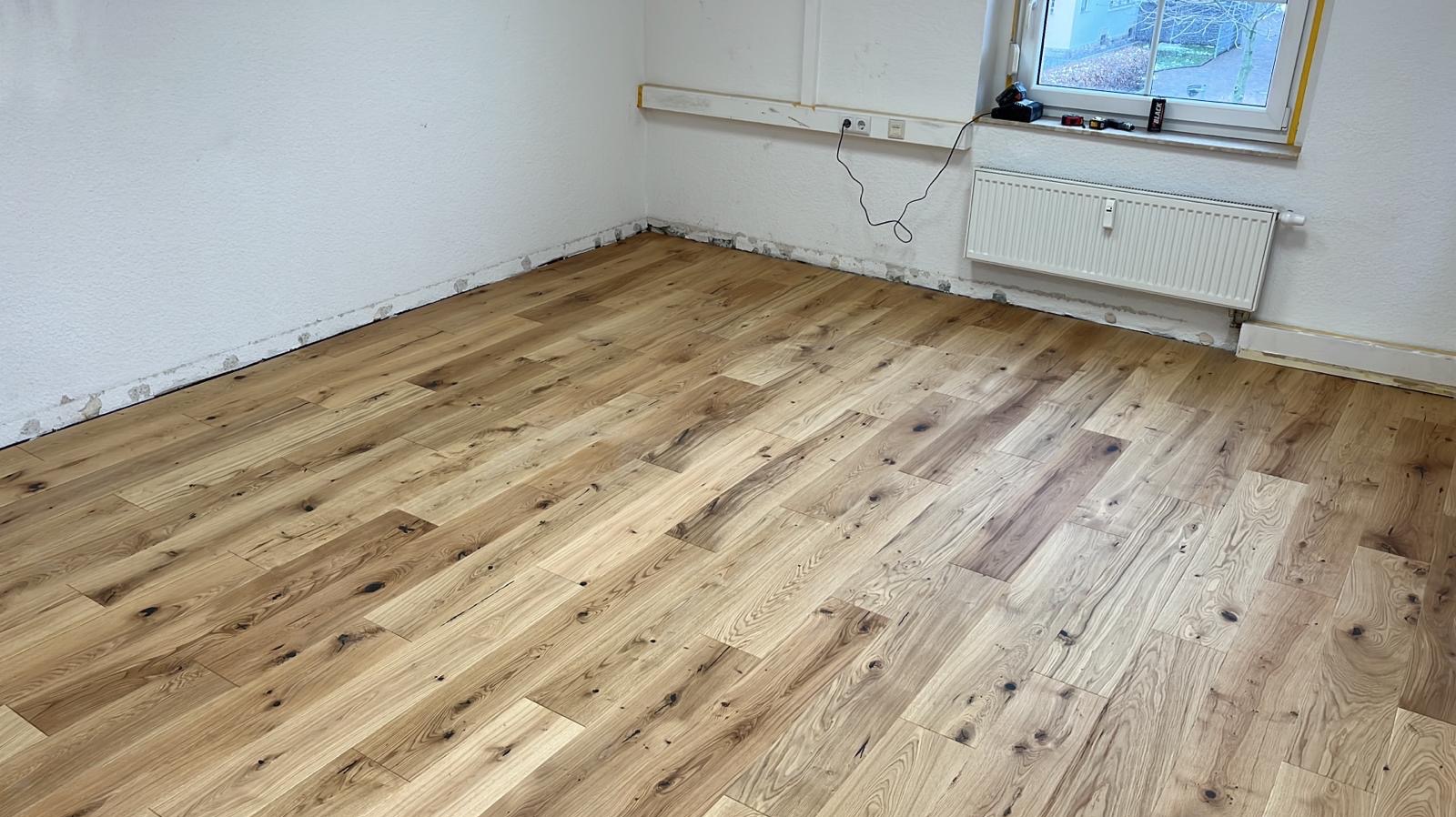 Ukończona podłoga z paneli drewnianych w jasnym odcieniu, ułożona w pokoju z białymi ścianami i grzejnikiem. Widoczne niedoskonałości przy ścianach przed listwami.