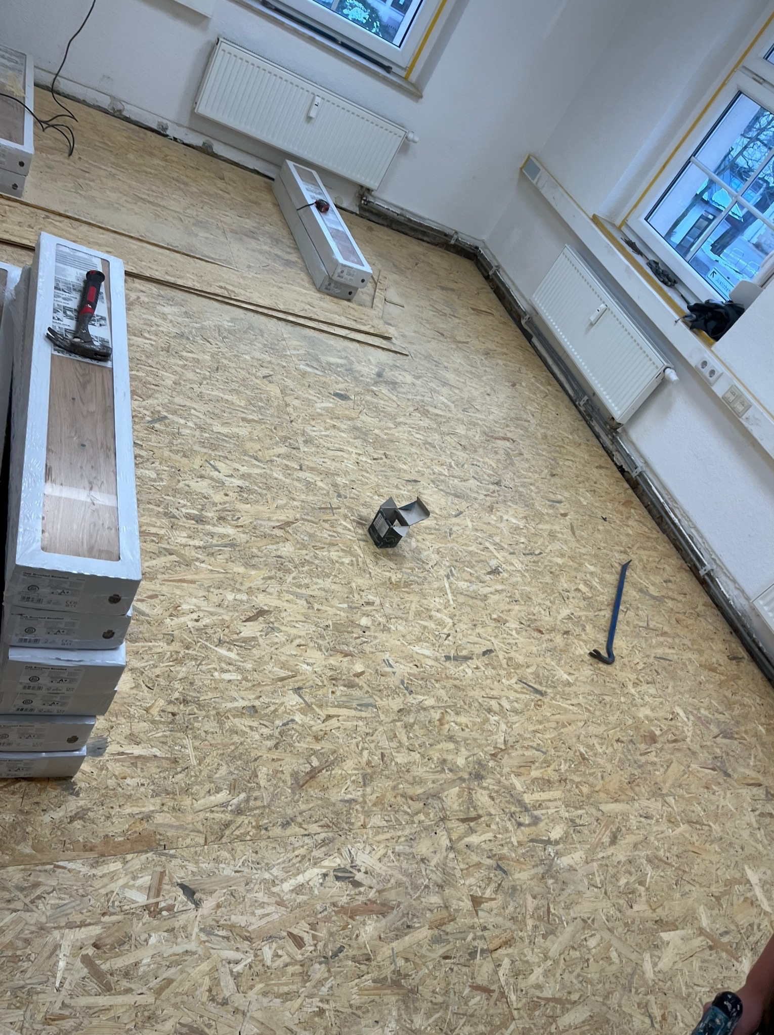 Przygotowanie do montażu paneli podłogowych: paczki paneli z młotkiem, podłoże OSB, listwy przypodłogowe i narzędzia w jasnym wnętrzu z grzejnikami.