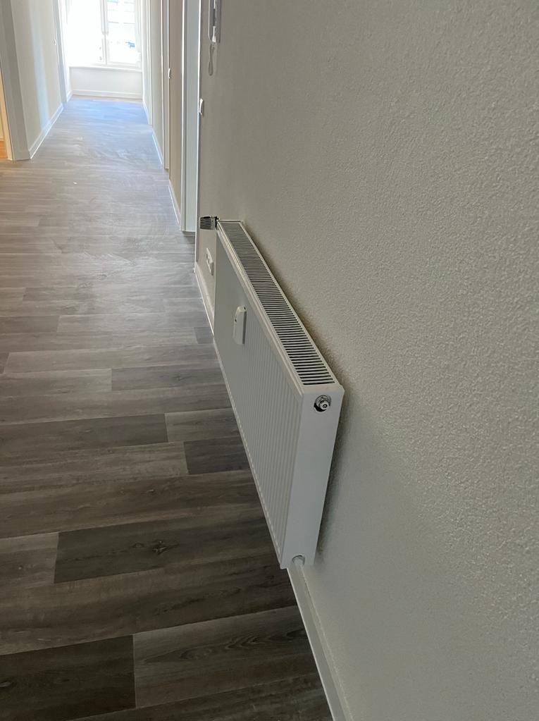 Biały grzejnik panelowy zamontowany na świeżo pomalowanej, jasnej ścianie w korytarzu z panelami podłogowymi. Widok z boku, perspektywa z dołu.