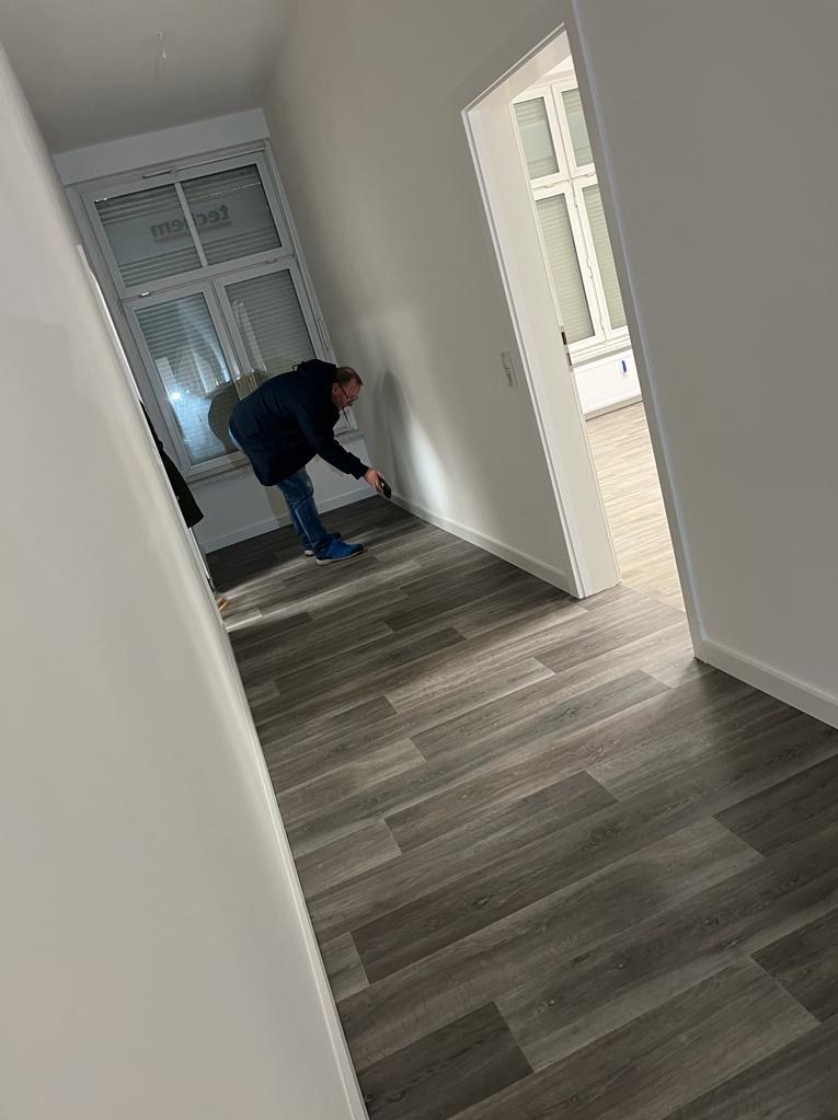 Montaż paneli podłogowych w korytarzu. Widoczny pracownik docinający panel przy ścianie. Szare panele imitujące drewno, białe ściany i okno z żaluzjami.