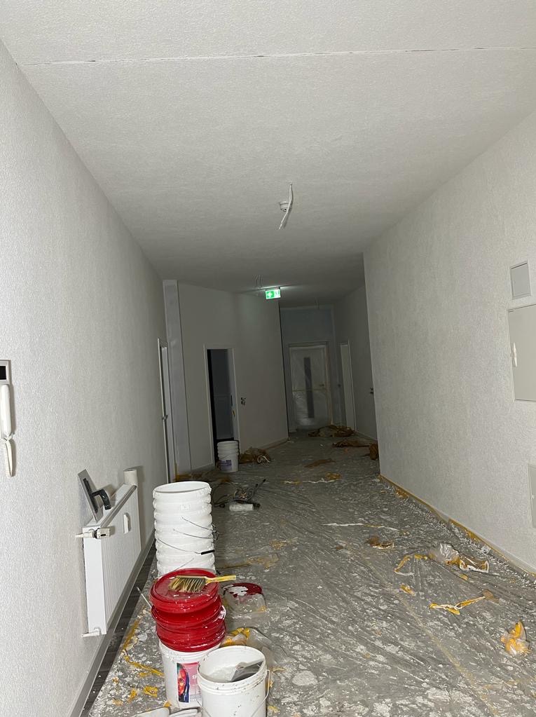 Biały korytarz po świeżym malowaniu, widoczne wiadra z farbą, folia ochronna na podłodze i narzędzia malarskie. Ściany gładkie, jednolite, z widocznym grzejnikiem.