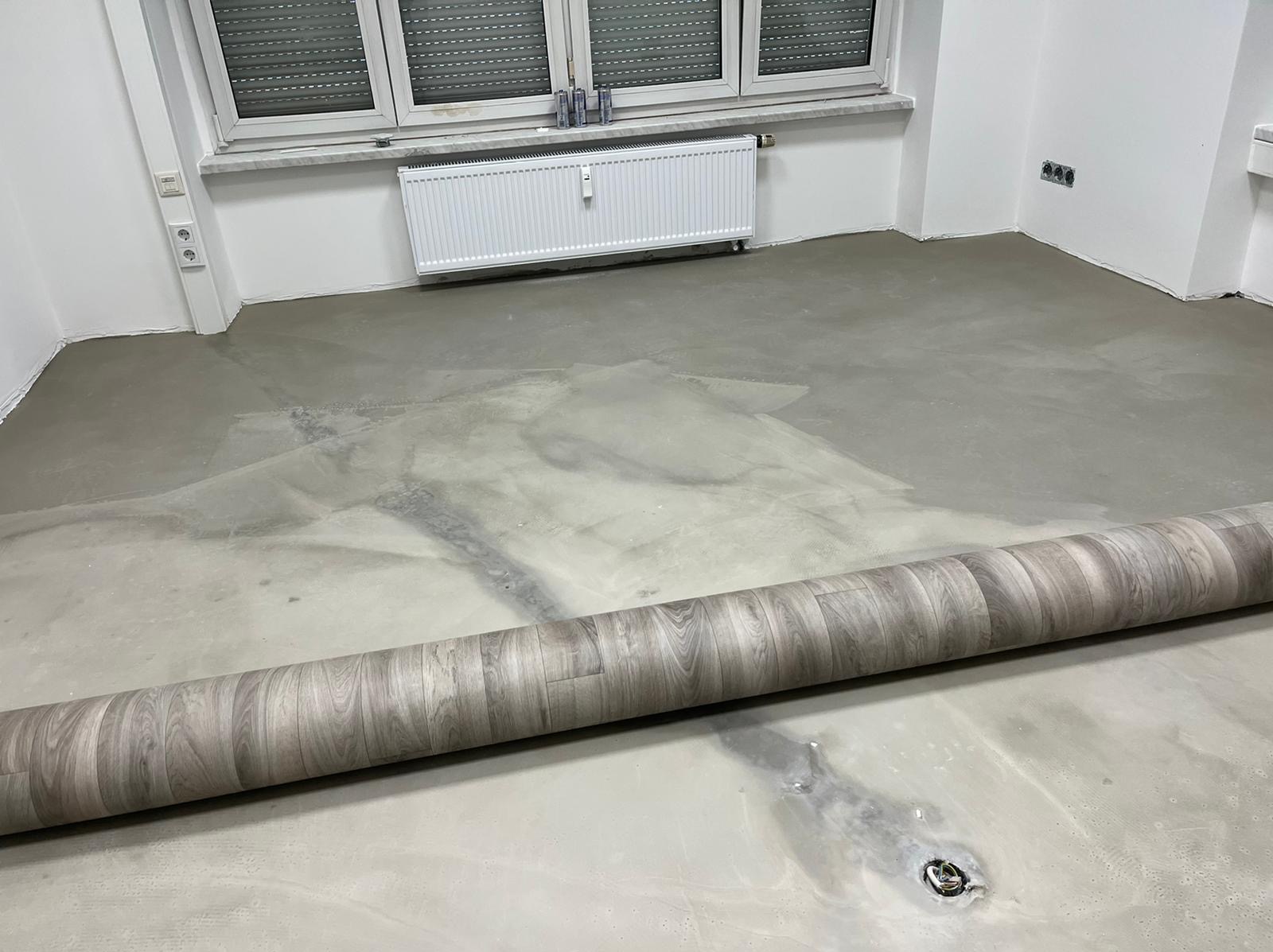 Przygotowanie do układania paneli: wyrównana, szara wylewka podłogowa w pokoju z oknem i grzejnikiem, zwinięty panel podłogowy z wzorem drewna w dolnej części kadru.