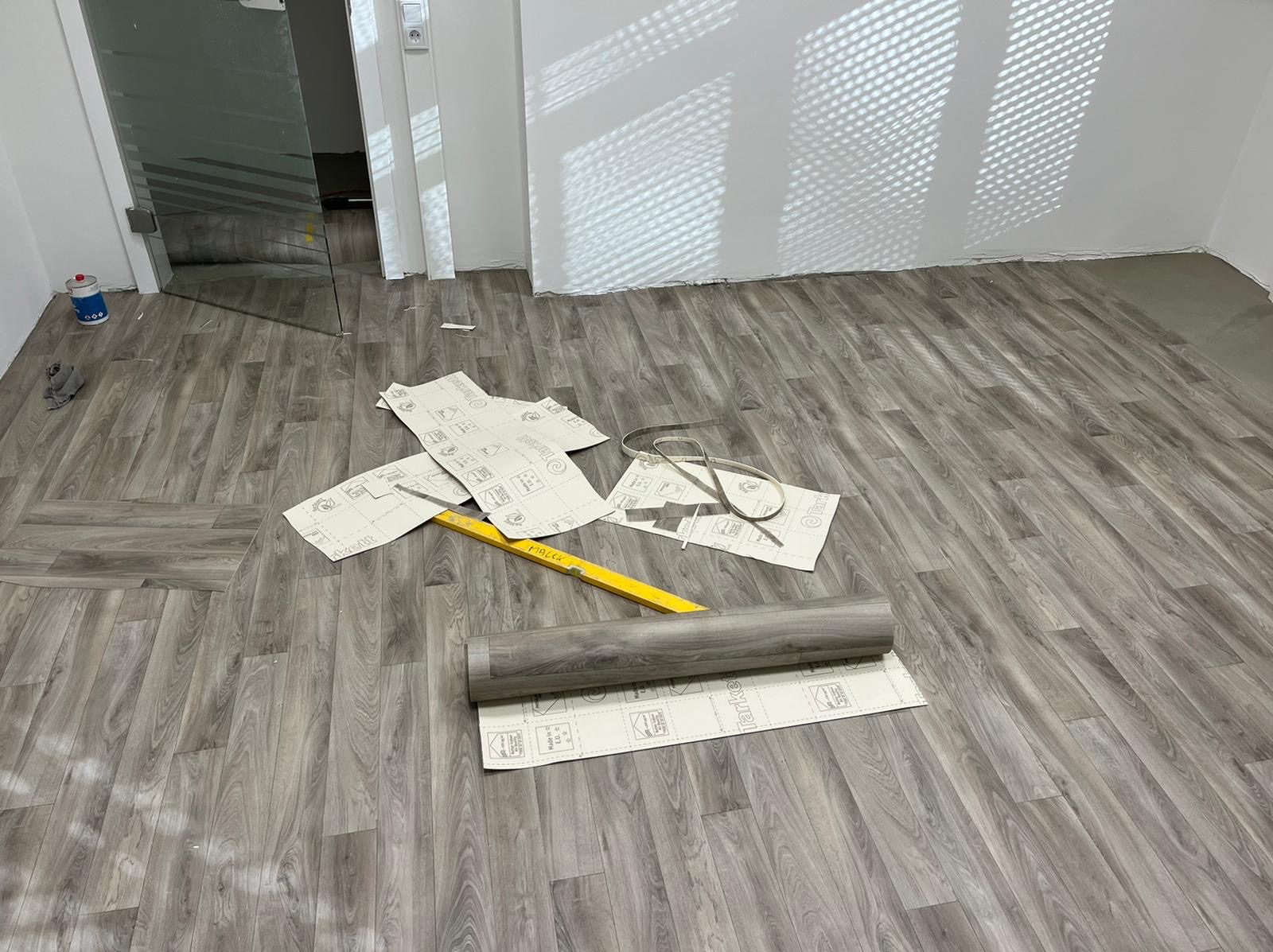 Jasnoszara podłoga z paneli, częściowo ułożona. Widoczny zwój paneli, poziomica i arkusze z instrukcją. W tle przeszklone drzwi i biała ściana z cieniem.