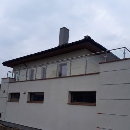 Steel Harmony Sp. z o.o. - Nowoczesna balustrada ze szkła i stali nierdzewnej na balkonie budynku. Minimalistyczny design, szare niebo w tle. Detal architektoniczny.