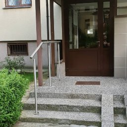 Steel Harmony Sp. z o.o. - Wejście do budynku z brązowymi drzwiami i metalową balustradą przy schodach. Widoczne płytki chodnikowe i zieleń. Nad drzwiami tabliczka z numerem lokalu.