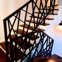 Steel Harmony Sp. z o.o. - Nowoczesne schody drewniane z czarną, geometryczną balustradą metalową. Widok z dołu, podkreślający unikalny design i solidne wykonanie.