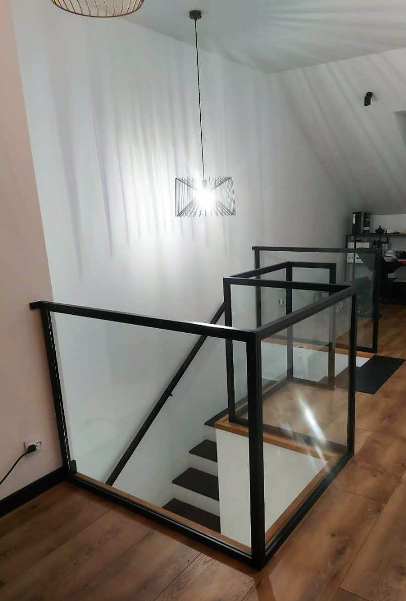Nowoczesne schody z czarną metalową balustradą i szklanymi panelami, oświetlone designerską lampą wiszącą. Widok z góry na drewniane schody.